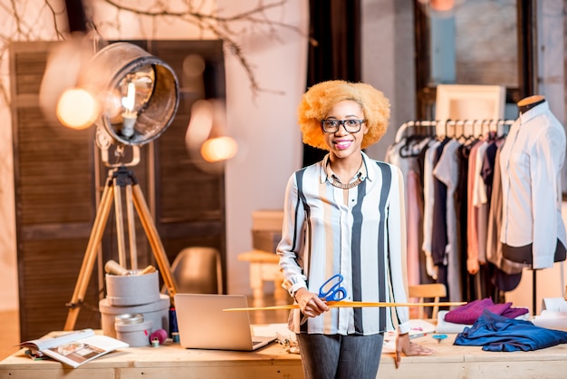 Portrait of a young african fashion designer standing with ruller at the office with different tailoring tools and clothes