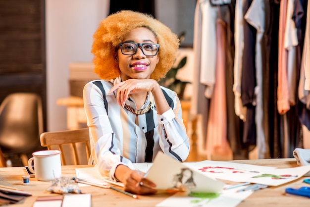 さまざまな仕立てツールと服を着てオフィスで働く若いアフリカのファッションデザイナーの肖像画