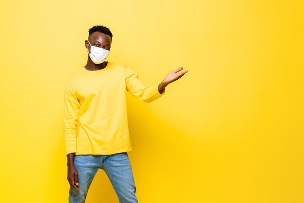 Portrait of young African man wearing face mask protecting from Corovavirus with hand opening to empty space aside in isolated yellow studio background