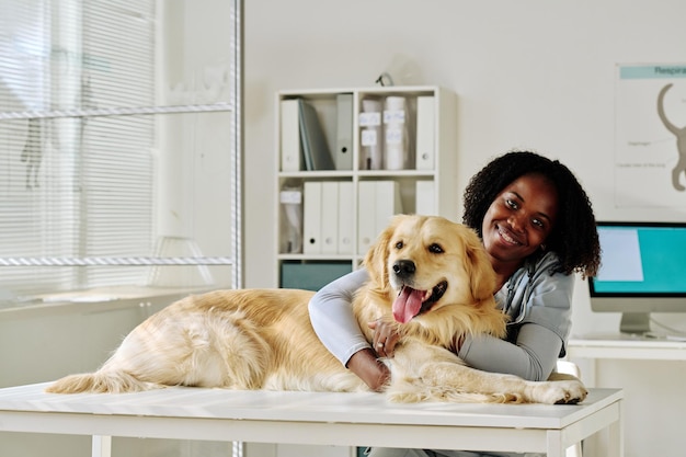 Ritratto di giovane medico veterinario africano che abbraccia il cane e sorride alla telecamera in ufficio