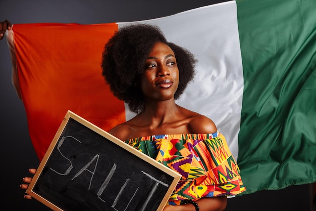 Photo portrait of young african woman student in national clothes holding a chalkboard with hello word on her native language before the ivory coast flag