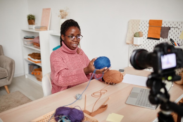 Portrait of young africanamerican woman knitting at home and recording video or livestream copy space