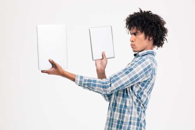 Portrait of a young afro american man choosing between tablet computer or small laptop computer isolated on a white wall