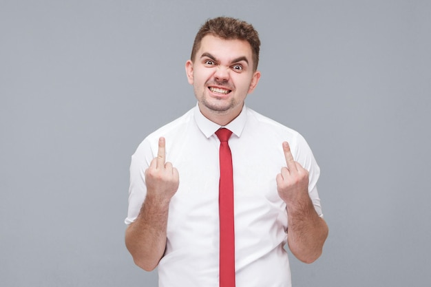 Portrait of young angry man in white shirt and tie standing\
with middle finger and angry face looking at camera indoor isolated\
on gray background