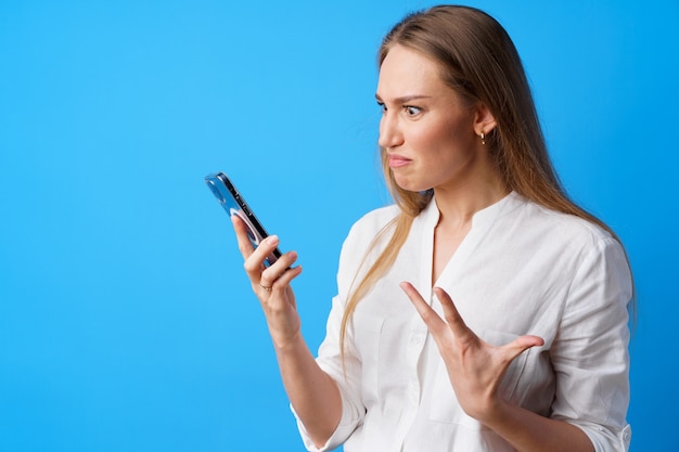 Portrait of young angry woman using her phone annoyed texting with someone blue background