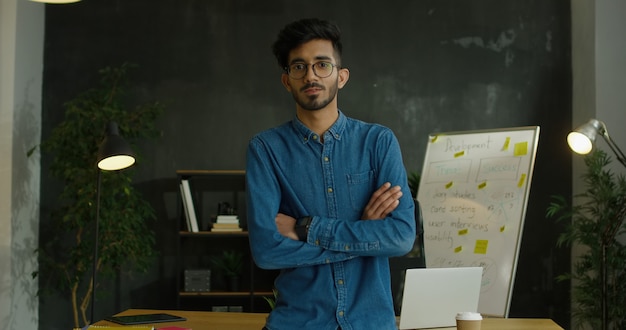 Photo portrait of young arab handsome guy in blue jeans shirt and glasses sitting on desk in office room, crossing hands and smiling to camera.