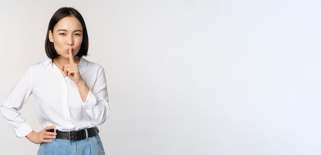 Portrait of young asian adult woman shushing say hush shh press finger to lips sharing secret dont speak taboo gesture standing over white background