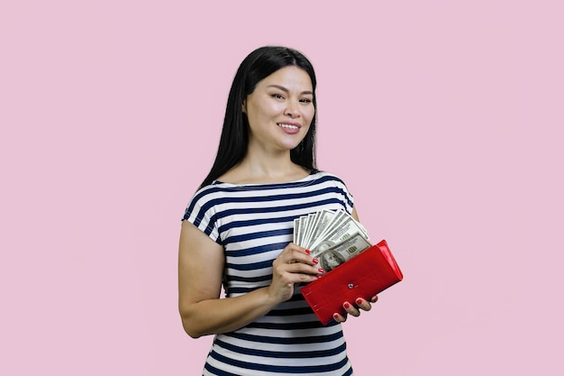 Portrait of a young asian brunette woman is putting dollars into a red wallet isolated on pink backg