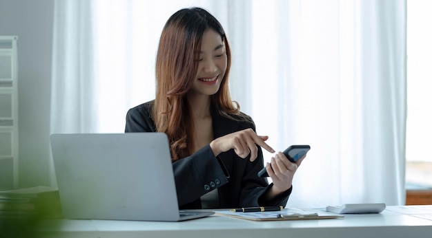 Портрет молодой азиатской бизнес-леди, использующей смартфон, пишет ноутбук в домашнем офисе. Красивая девушка за настольным компьютером принимает заказ на дом.