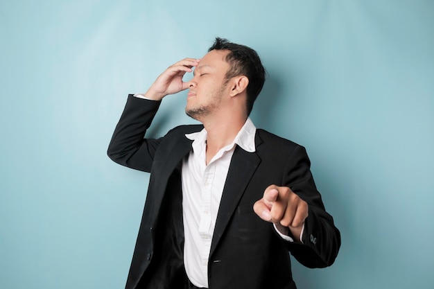 Portrait of young Asian businessman in black suit pointing index finger at camera posing isolated over blue background Cheerful smiling guy picking choosing and indicating