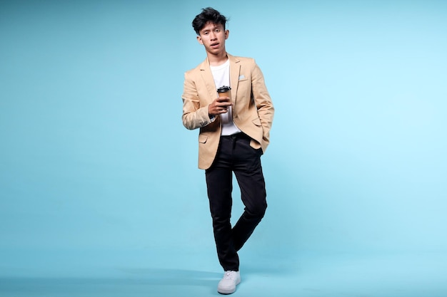 Portrait of young Asian businessman holding coffee cup and looking at camera isolated on blue backgr