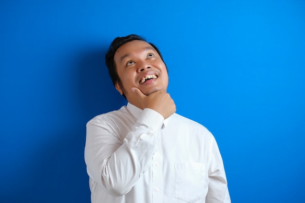 Portrait of young Asian businessman thinking something, smiling happily for having good idea, find solution for a problem, against blue background with copy space