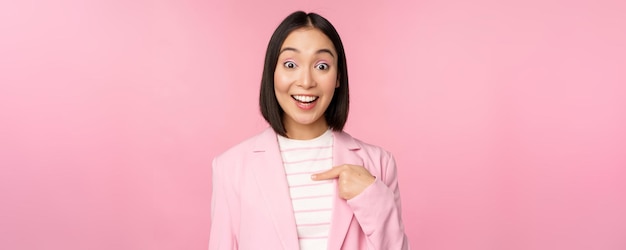 Ritratto di giovane donna d'affari asiatica con espressione faccia eccitata sorpresa che punta il dito contro se stessa in piedi in tuta su sfondo rosa
