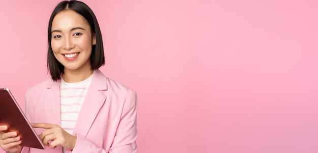 若いアジアの企業の女性のオフィスの女性の肖像画は、ピンクの背景に対して笑顔でプロのポーズをとってスーツを着てデジタルタブレットを持っています