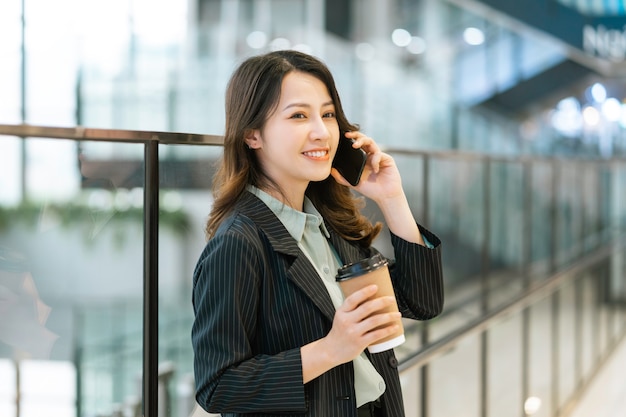 Портрет молодой азиатской женщины-директора, стоящей, пьющей кофе и слушающей телефон