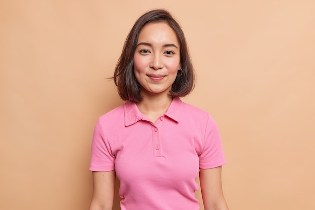 Portrait of young Asian female model with natural beauty dark hair rouge cheeks healthy skin looks happily at front wears casual pink t shirt isolated over beige wall