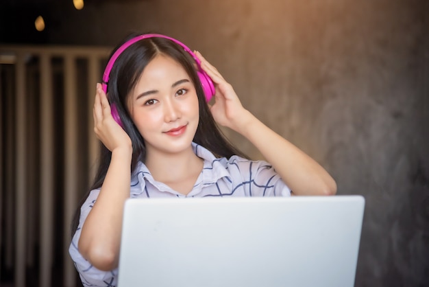 Ritratto delle cuffie d'uso di giovane felicità asiatica facendo uso del computer portatile a casa per lavorare