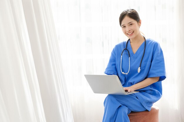 Photo portrait of a young asian happy nurse wearing medical scrubs looking at the camera with a stethoscope and using a laptop while sitting with a white curtain in the background image with copy space
