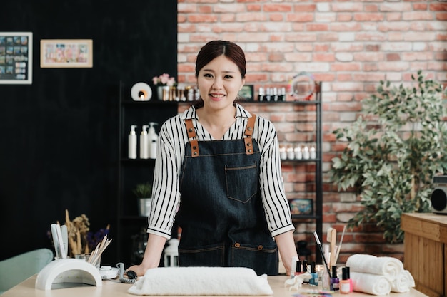 Portrait of young asian korean girl owner of beauty salon for nail care and hands standing in studio. attractive woman beautician face camera smiling at working table. manicurist in apron in workshop