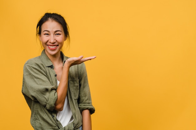 Portrait of young Asian lady smiling with cheerful expression, shows something amazing at blank space in casual cloth  isolated over yellow wall. Facial expression concept.