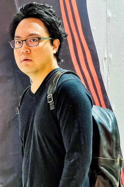 Photo portrait of young asian man in eyeglasses and backpack standing against patterned wall
