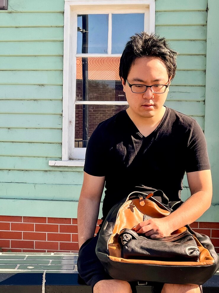 Photo 건물 외관 앞 의 보행자 길 에 앉아 있는 안경 을 입은 젊은 아시아인 의 초상화