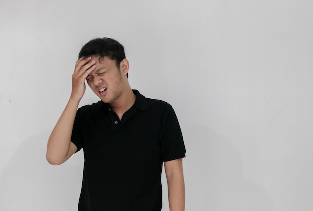 Portrait of young asian man isolated on grey background suffering from severe headache pressing fingers to temples closing eyes to relieve pain with helpless face expression