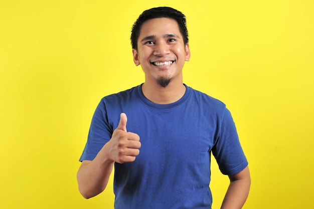 Portrait of young Asian man laughing with showing thumbs up at camera, isolated on yellow