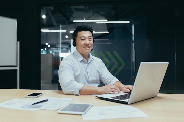 Portrait of a young asian man sitting in the office at a desk in a headset working on a laptop he