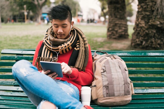 屋外のベンチに座ってイヤホンで彼のデジタルタブレットを使用して若いアジア人の肖像画。