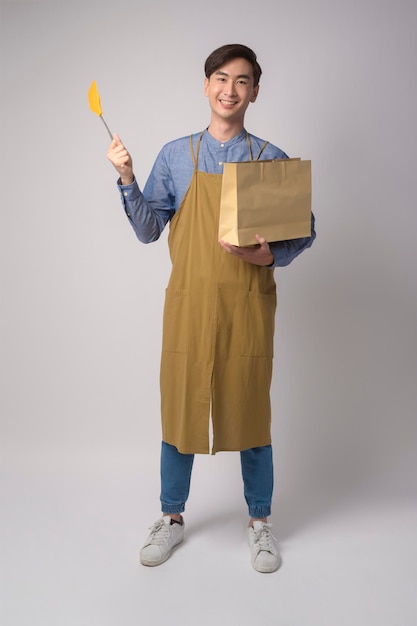 Portrait of young asian man wearing apron holding paper bag and turner over white background studio