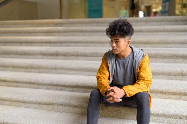 Photo portrait of young asian man with curly hair sitting on staircase in the city