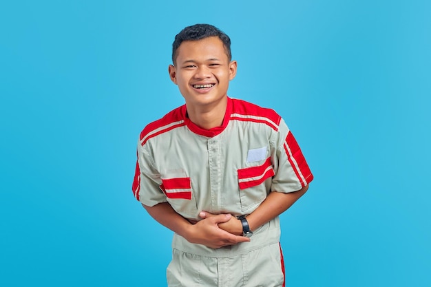 Portrait of young Asian mechanic showing loud laughter with hands on stomach over blue background