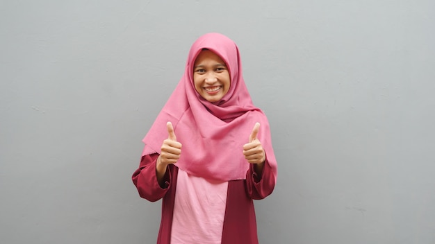 Portrait of young Asian muslim lady wearing hijab shows thumbs up gesture, smiling happy face, approved OK sign