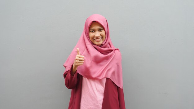 Portrait of young Asian muslim lady wearing hijab shows thumbs up gesture, smiling happy face, approved OK sign