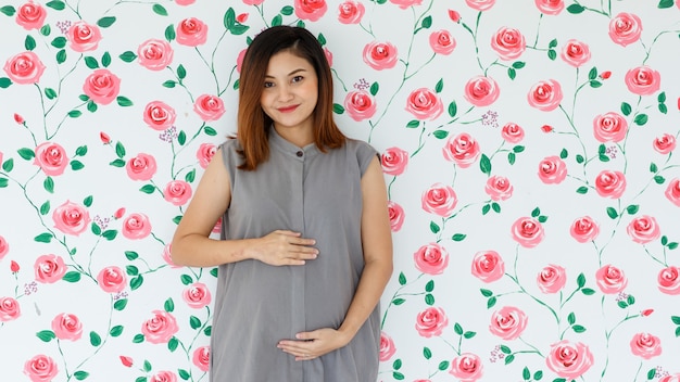 Portrait of young Asian pregnant woman smiling standing touching her belly looking at a camera over white roses pattern background. Healthy and happy lady concept.