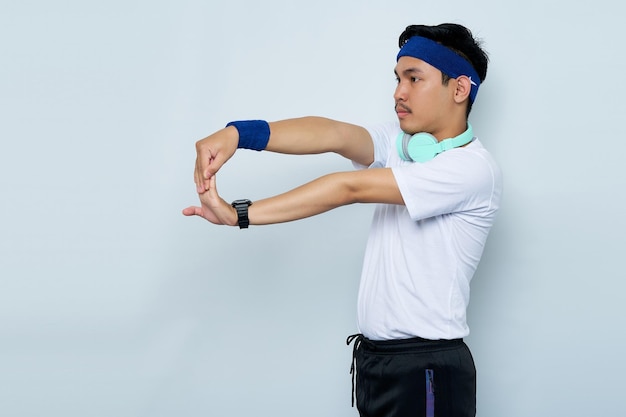 Portrait of young Asian sportman in blue headband and sportswear white tshirt with headphones working out doing stretching exercising looking aside isolated on white backgroundWorkout sport concept