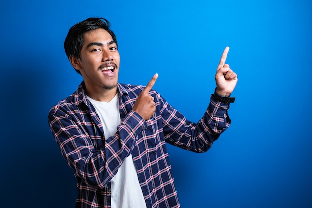 Portrait of young Asian student wearing shirt smiling and pointing to presenting something on his side, against blue background with copy space
