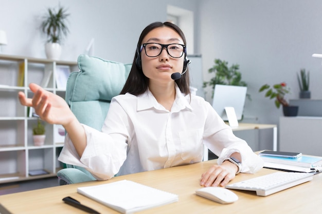 Portrait of a young asian woman in glasses and headphones with a microphone sitting in the office at