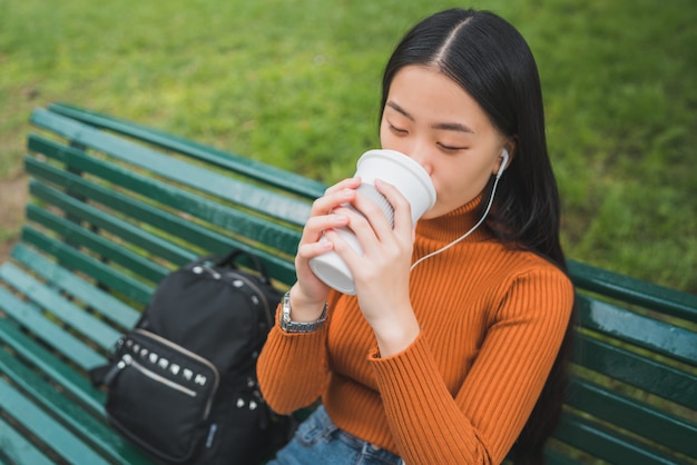 이어폰으로 음악을 듣고 야외 공원에서 커피 한 잔을 마시는 젊은 아시아 여자의 초상화.