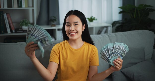 Portrait of young Asian woman showing dollar at camera and smiling on sofa in the living room at home