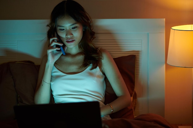 Portrait of young Asian woman sitting on bed and calling coworker to discuss new release