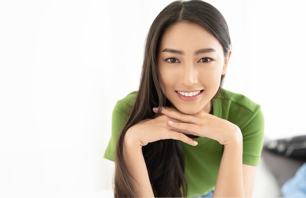 Portrait of young Asian woman smiling friendly with hands on chin and looking at camera in living room Woman's face closeup Concept woman lifestyle