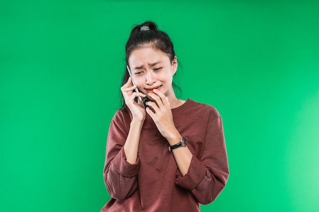 緑の背景に彼の手で彼のあごを保持しながら、泣いて、ショックを受けた表情で電話で話している肖像画の若いアジアの女性