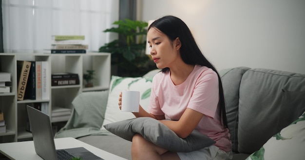 Портрет молодой азиатской женщины, думающей и использующей ноутбук во время питья кофе на диване дома