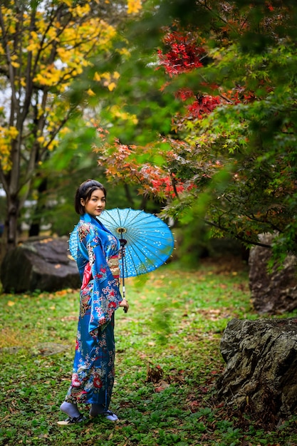 日本の紅葉シーズンに公園に立っている日本の青い着物のファッションの歴史と傘を身に着けている若いアジアの女性の肖像画