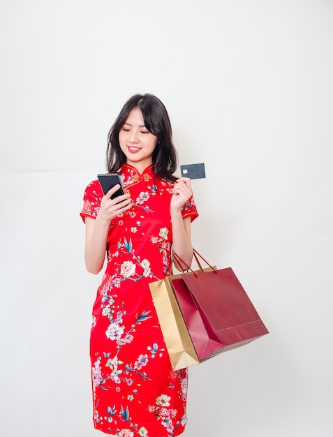 Ritratto di una giovane donna asiatica che indossa il tradizionale abito cheongsam qipao in possesso di borse della spesa per lo smartphone e mostra la carta di credito isolata su sfondo bianco