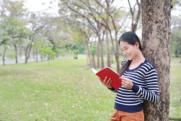 Женщина портрета молодая азиатская с положением книги постным против ствола дерева в парке внешнем.