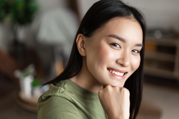 Portrait of young asian woman with clean glowing skin looking and smiling at camera sitting at home ...