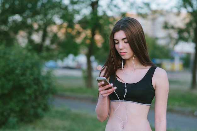 Portrait of a young athletic girl listening to music on headphones and use the phone in the park. Sports Portrait beautiful girl in headphones outdoors.